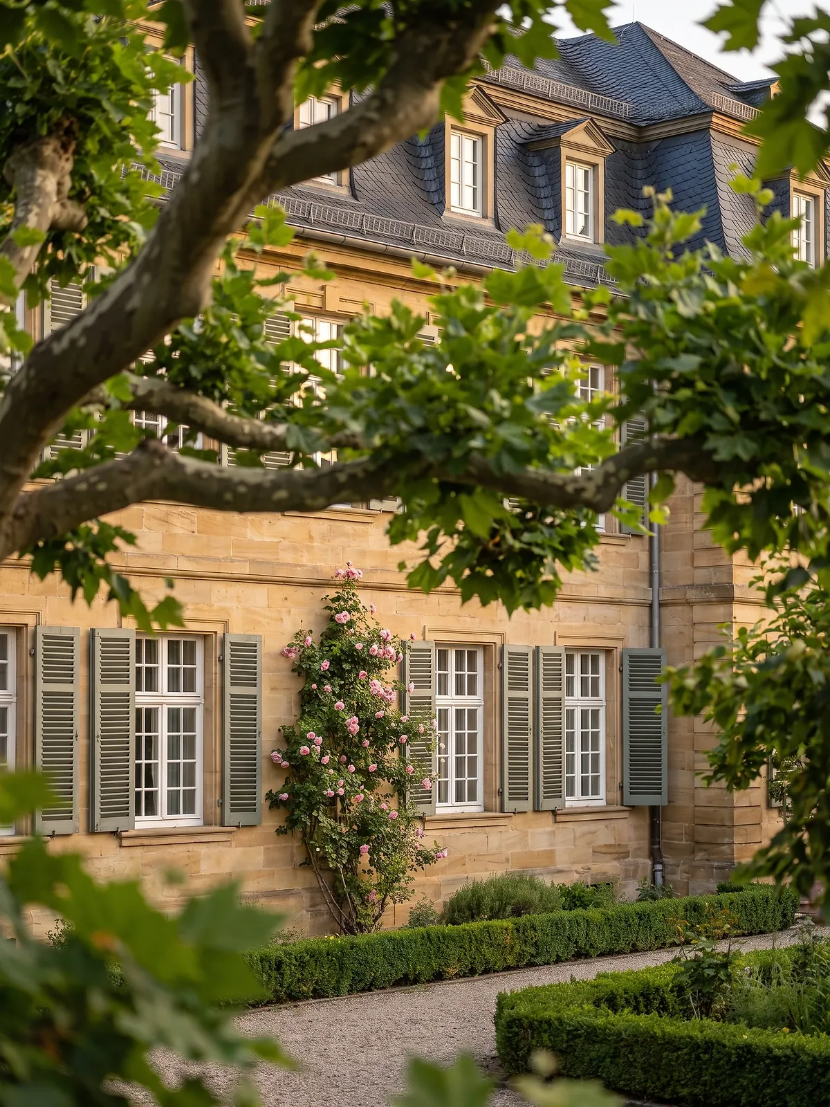 Sandsteinfassade mit grünen Fensterläden und Kletterrose hinter dem Laub einer Platane (KI-generiertes Symbolbild)